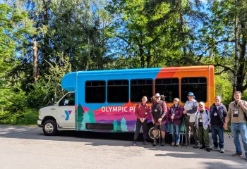 A group of Senior adults gathered around the Y van in the woods.
