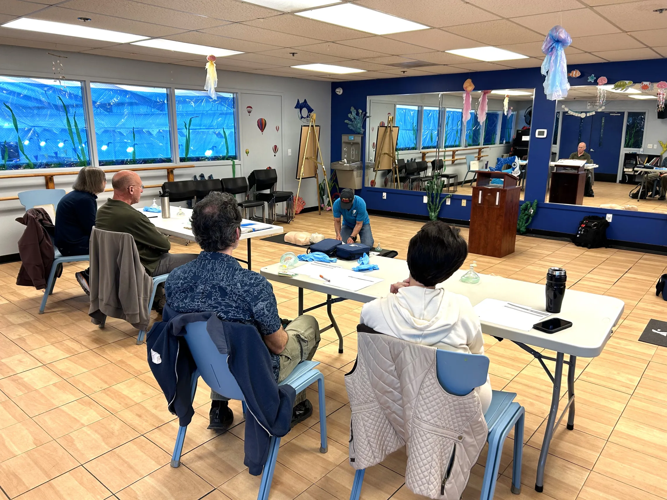 Classroom full of adults sitting watching a CPR demonstration by the instructor