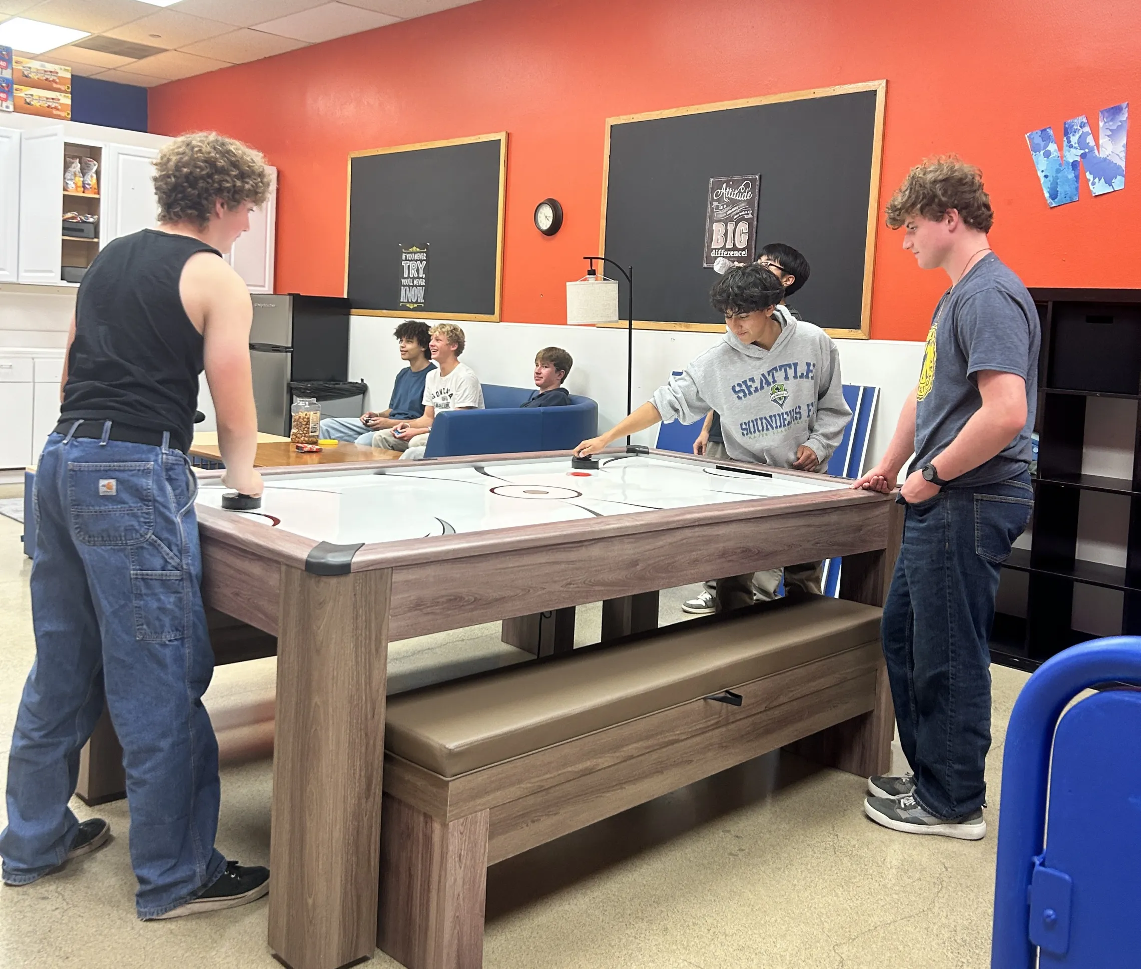 A group of teens playing air hockey and sitting on a couch playing video games.