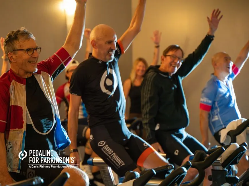 Adult men and women using stationary bikes with one arm raised.