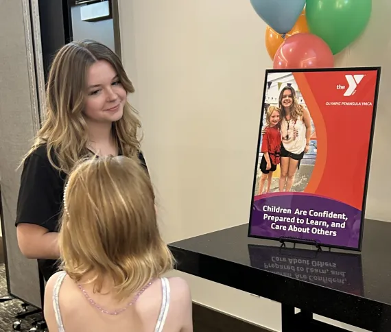 A young woman talking to a young girl while they look on at a poster of the two of them