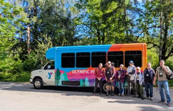 A group of Senior adults gathered around the Y van in the woods.